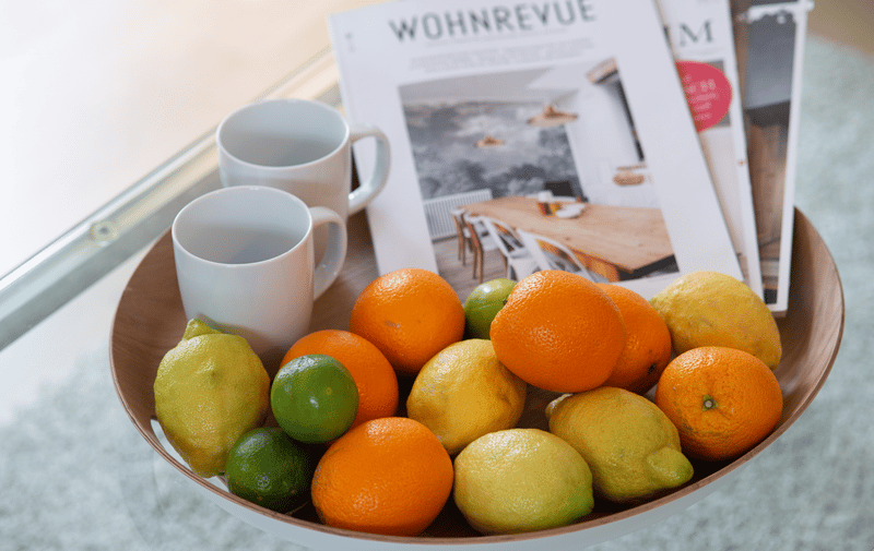 A brown bowl filled with citrus fruits, such as lemons, limes, and oranges, along with two white coffee mugs and a magazine placed on a glass table.