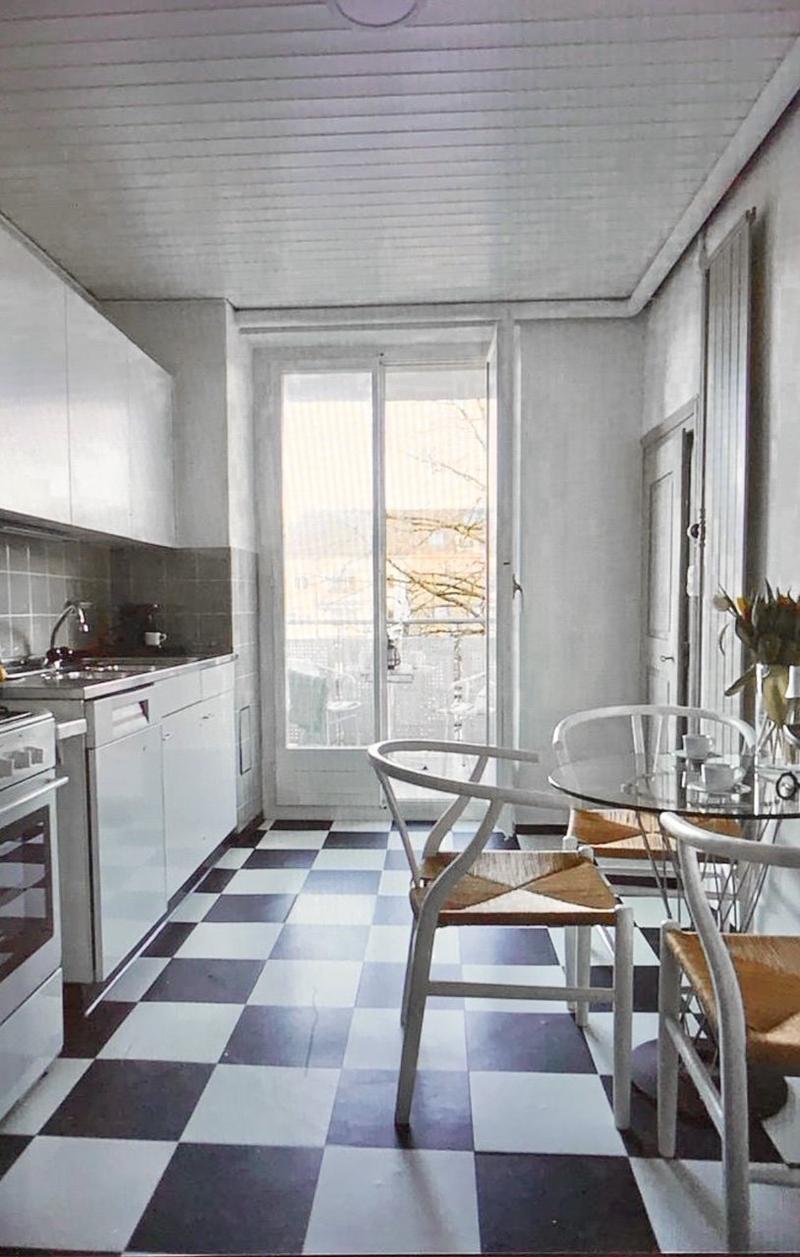 black and white checkered floor, white walls, white and grey kitchen cabinets, sink, oven, dishwasher, table and chairs, glass door leading to balcony
