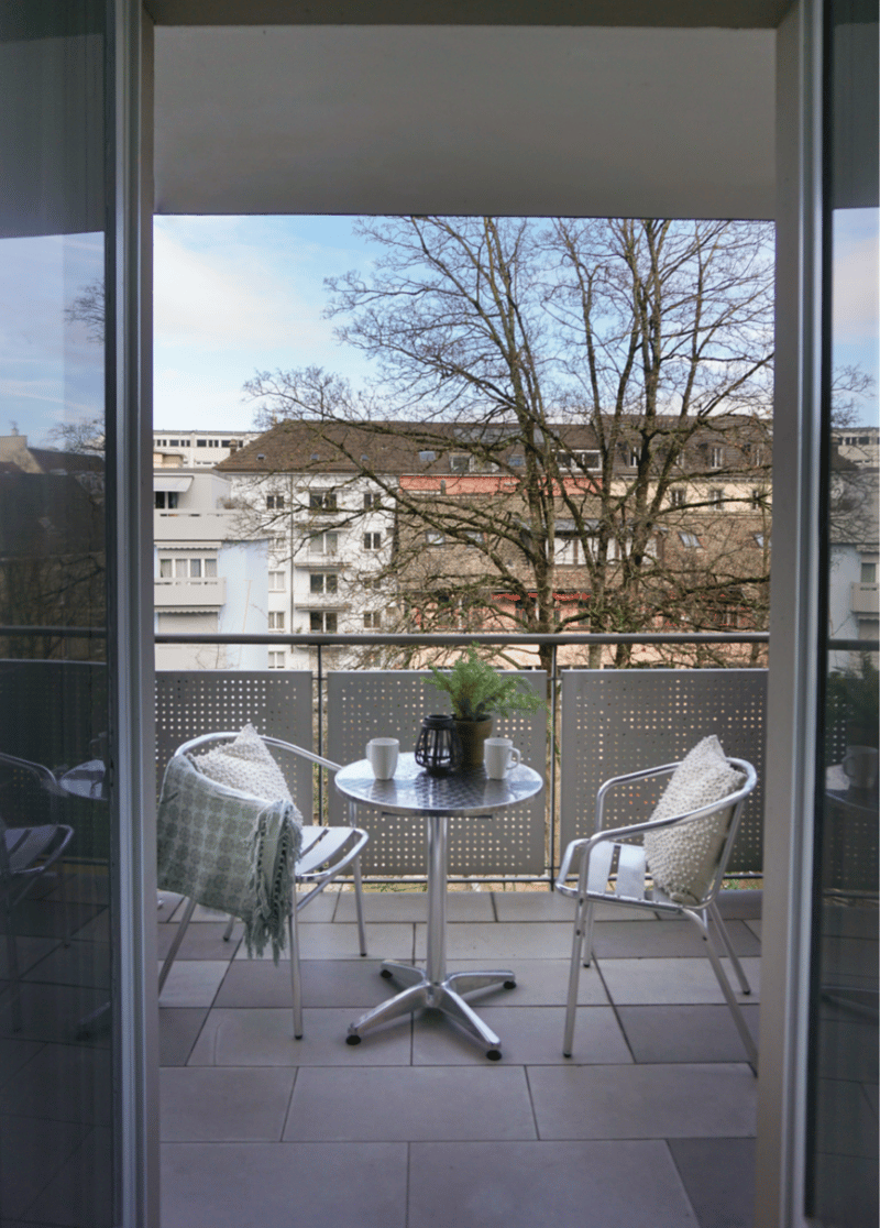 Modern balcony with table and chairs, potted plant, and metal railings. The balcony has a view of the surrounding buildings and trees.