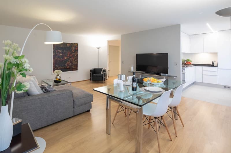 modern living room with gray couch, glass table, white chairs, TV, artwork, floor lamp, flowers, kitchen area