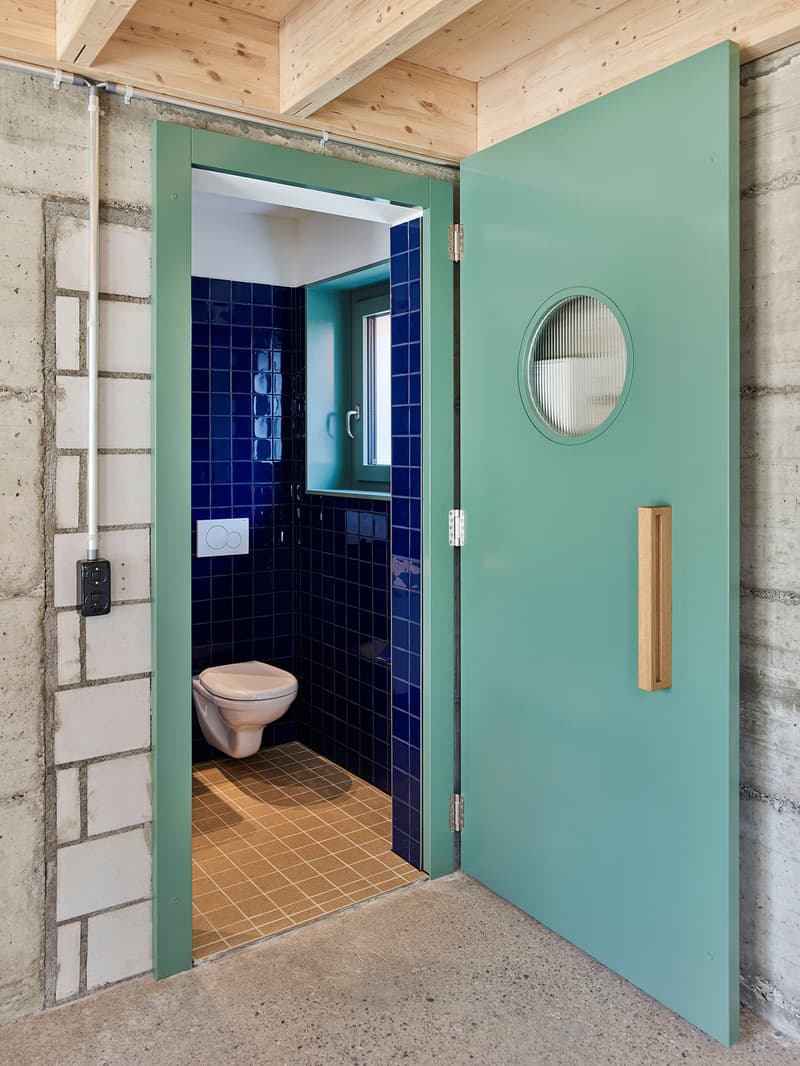 The image shows a bathroom with a wooden ceiling, concrete walls, and a teal-colored door frame. The bathroom features blue tile walls, a white toilet, and a round mirror on the wall. The floor is tiled with a light-colored tile.