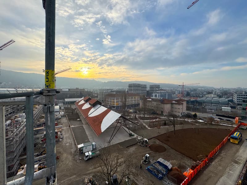 The image shows a construction site with various buildings and cranes in the background. The sky is filled with clouds and the sun is setting, creating a warm, golden glow. There are several construction vehicles and equipment visible on the site.