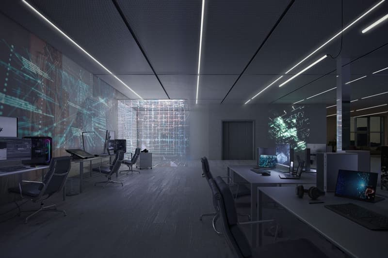 Large open office space with modern, high-tech design elements. Includes multiple desks and workstations, computers, and a large projection screen or display on the wall displaying digital graphics and data visualizations.