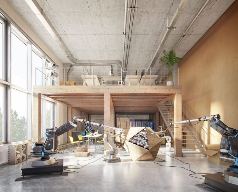 Large open-concept workspace with concrete ceiling, wooden beams, and large windows. The space is filled with various equipment, tools, and materials, suggesting it is used as a workshop or studio. There is a mezzanine level above, and the overall atmosph
