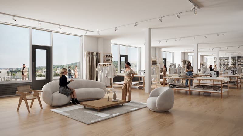 Large open-concept retail space with high ceilings, large windows providing a view, hardwood floors, and modern furniture and decor. The space includes a seating area with a large gray sofa and coffee table, as well as clothing racks and displays througho