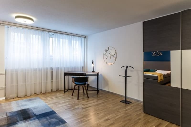 Spacious bedroom with large windows, hardwood flooring, a desk with a chair, and a wardrobe cabinet. The room has a modern and minimalist design.