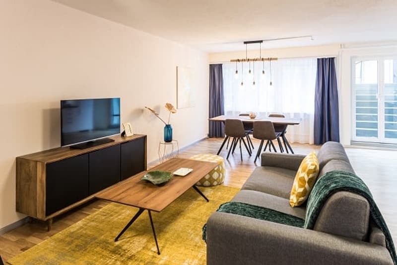 Open-concept living room and dining area, hardwood floors, large windows, gray sofa, wooden coffee table, TV stand, dining table with chairs, pendant lighting