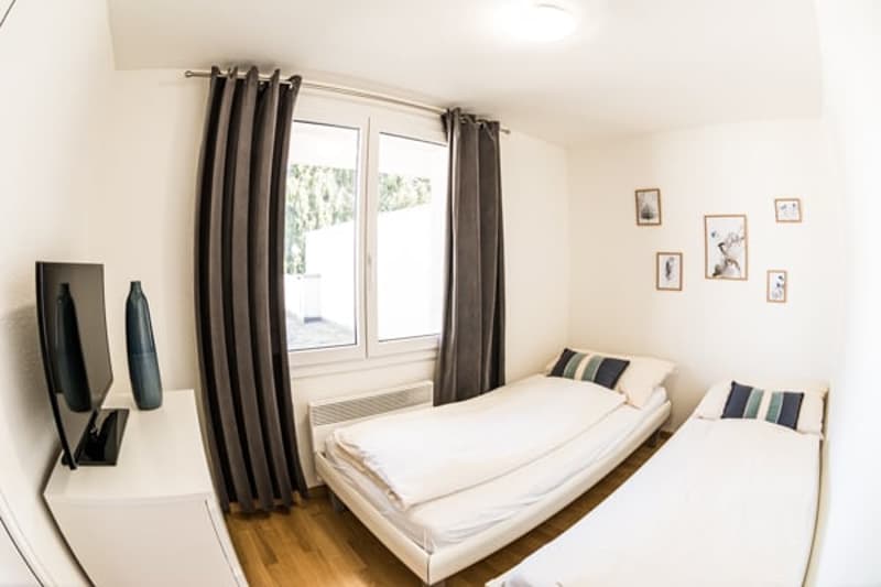 Two single beds, flatscreen TV on white side table, grey curtains, wooden floor, picture frames
