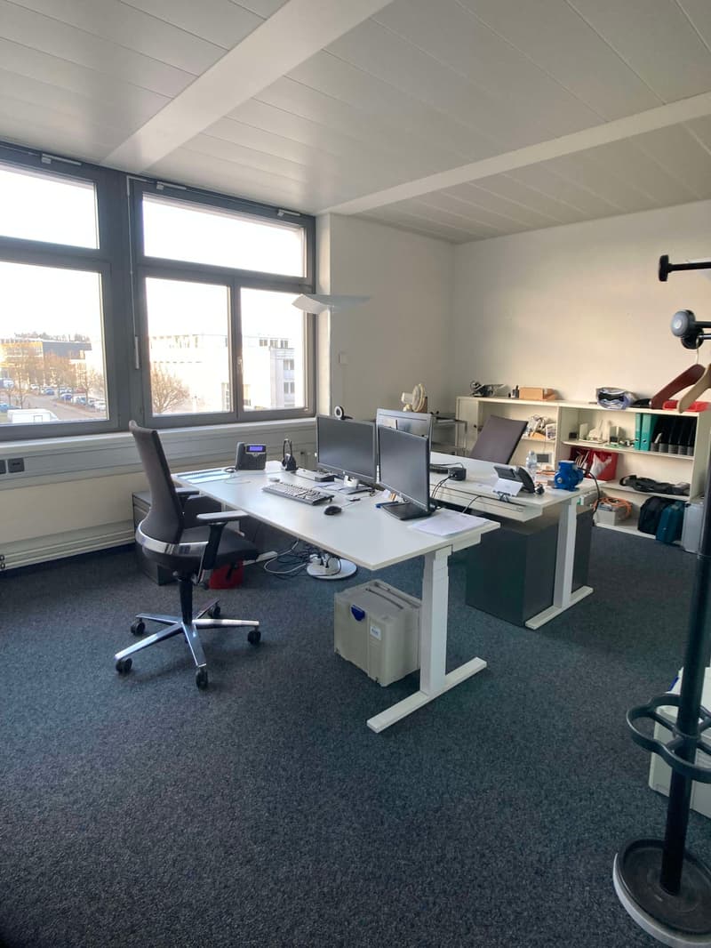 The image shows a modern office space with large windows, a white ceiling, and gray carpeted flooring. There are two desks with computers, monitors, and other office equipment. The room also contains shelves and storage units for various items. The overal