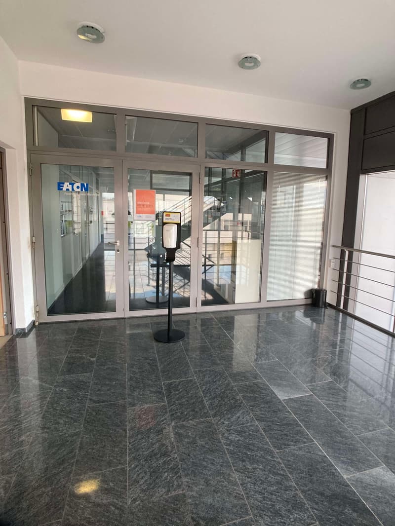The image shows the interior of an office lobby or entrance area. The space features a tiled floor with a gray marble-like pattern, and large glass windows and doors that allow natural light to enter. There is a security check-in station with a podium in