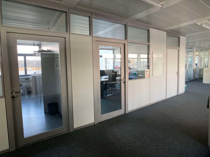The image shows an office space with glass partitions and doors, creating separate rooms or workspaces. The walls are light-colored, and the floor is covered with dark carpeting. There are desks, chairs, and other office equipment visible through the glas