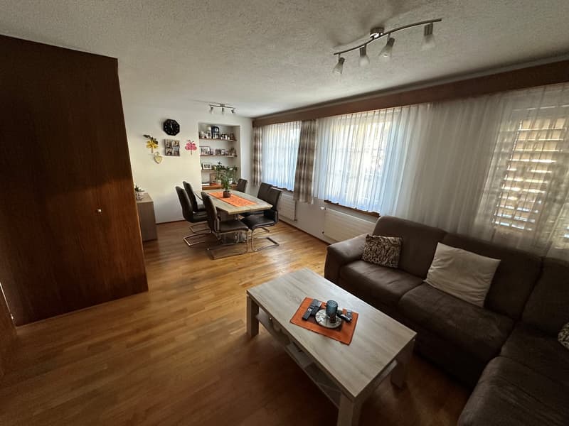Dining room: wooden floor, dining table with 6 chairs, shelves with decorative items, white curtains, kitchen area.