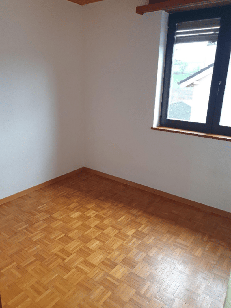 The image shows an empty bedroom with white walls and a wooden parquet floor. There is a window with a black frame providing natural light to the room.