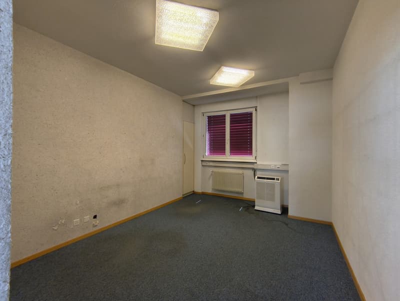 Spacious bedroom with gray carpeted floor, white walls, and two ceiling lights. The room includes a window with blinds and an air conditioning unit.