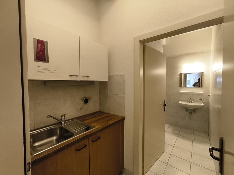 The image shows a small studio apartment with a kitchenette and a bathroom. The kitchenette has a sink, cabinets, and a wooden countertop. The bathroom is visible through the open doorway and has a sink and mirror. The room appears to be clean and well-ma
