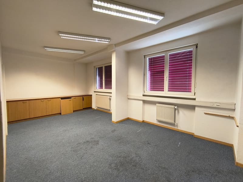 The image shows a spacious office space with beige walls, gray carpeted floor, and wooden cabinets along the wall. The room is well-lit with fluorescent lighting fixtures on the ceiling. There are two large windows with purple blinds, providing natural li