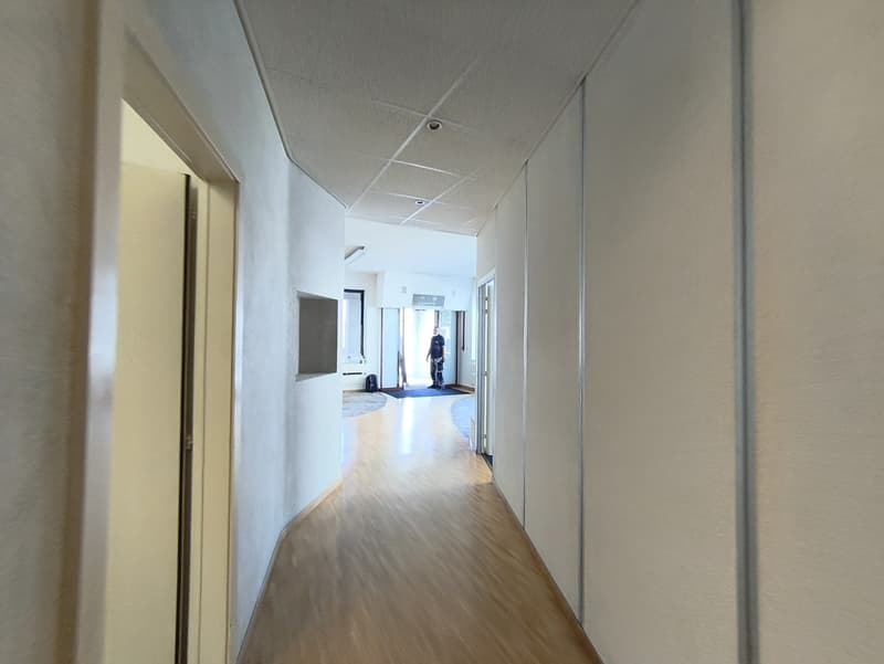 The image shows a long, well-lit hallway with a white ceiling and walls. The floor is made of hardwood or a similar material. There is a person visible in the distance, walking down the hallway. The hallway appears to be part of an office or commercial bu