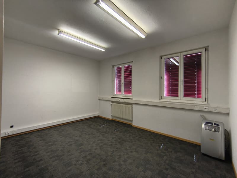 Empty room with gray walls, gray carpeted floor, two windows with red blinds, and fluorescent lighting fixtures on the ceiling. There is also a portable air conditioning unit in the room.