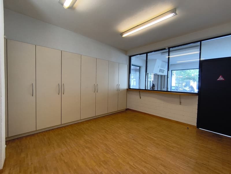 This image shows a large, empty room with white walls and a wooden floor. The room has several built-in cabinets or storage units along one wall, and a large window that provides natural light. The room appears to be a multipurpose space, possibly used fo