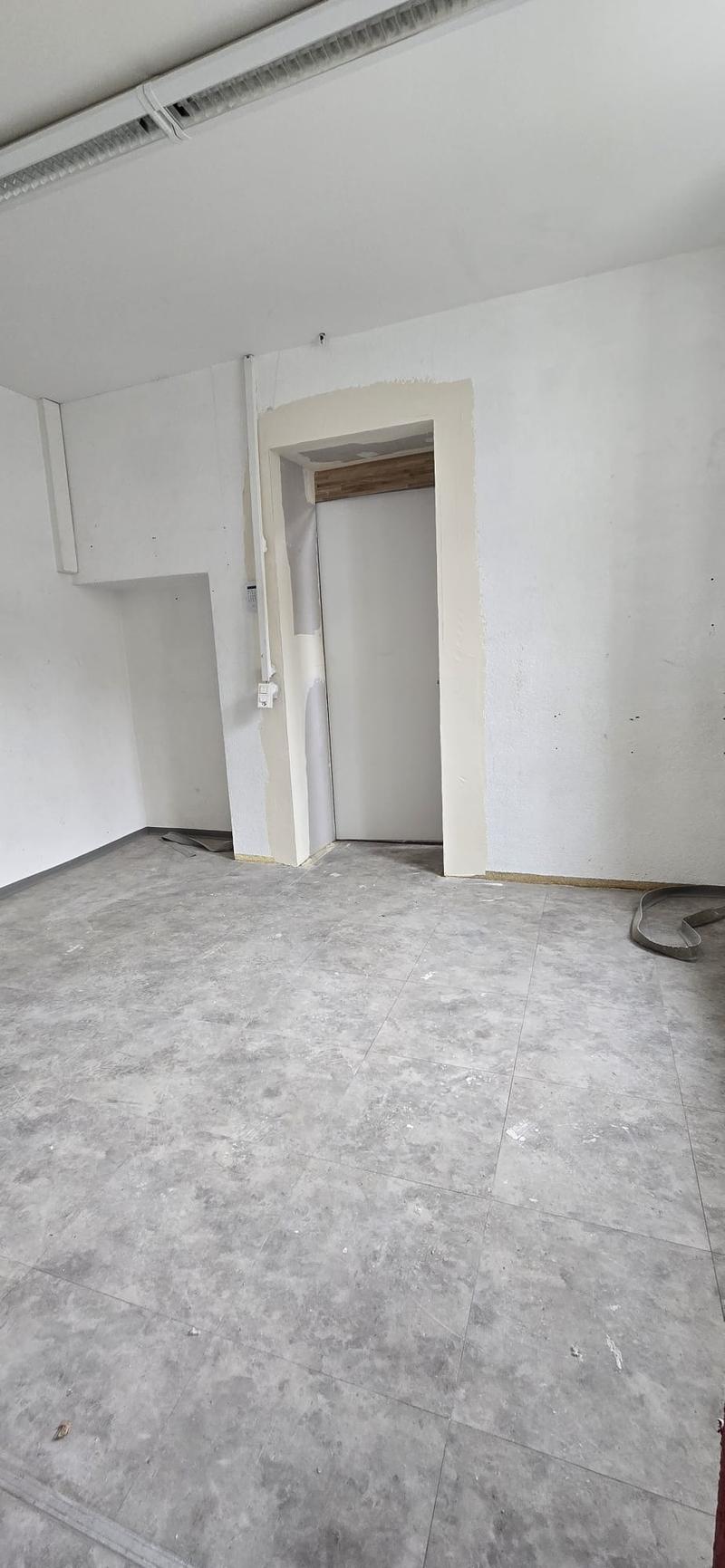 Empty room with large windows, white walls, concrete floor, white ceiling, exit door.