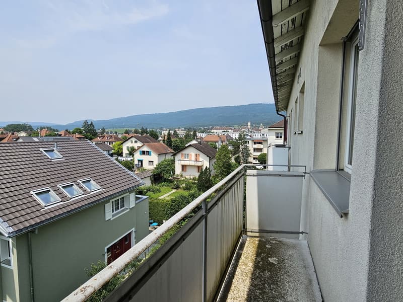 The image shows a balcony or terrace overlooking a residential neighborhood. The balcony has a railing and there are several houses and apartment buildings visible in the distance, with some trees and greenery in the foreground.