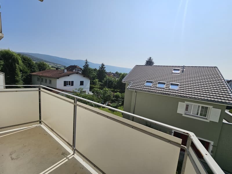 The image shows a residential building with a balcony or terrace. The building has a green exterior and a tiled roof. There are trees and other buildings visible in the background, suggesting a suburban or residential area.