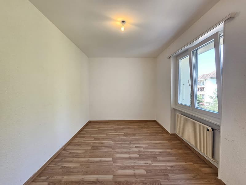 This image shows a spacious bedroom with white walls and a hardwood floor. The room has a large window that provides natural light and a view of the surrounding area. The room appears to be empty, with no furniture or decor visible.