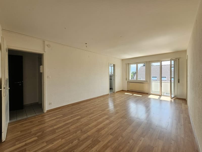 Spacious living room with hardwood flooring, white walls, and large windows leading to a balcony or terrace. The room appears to be well-lit and has an open layout.
