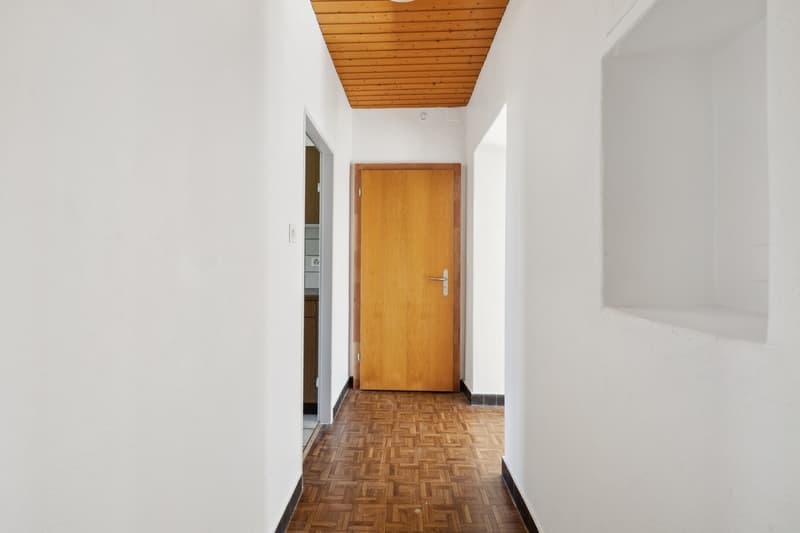 The image shows a hallway with a wooden ceiling, white walls, and a wooden door. The floor is made of parquet tiles in a geometric pattern.