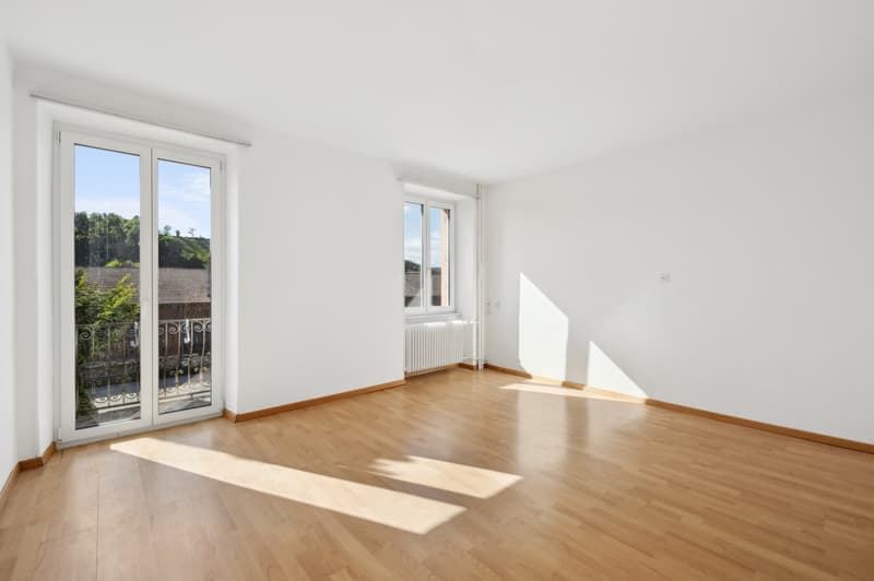 Spacious living room with large windows, hardwood flooring, and access to a balcony or terrace overlooking the surrounding greenery.