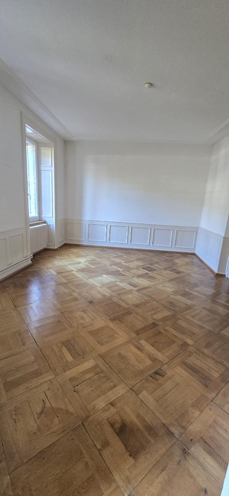 Spacious room with white walls, wooden parquet flooring in a herringbone pattern, and a single light fixture on the ceiling.