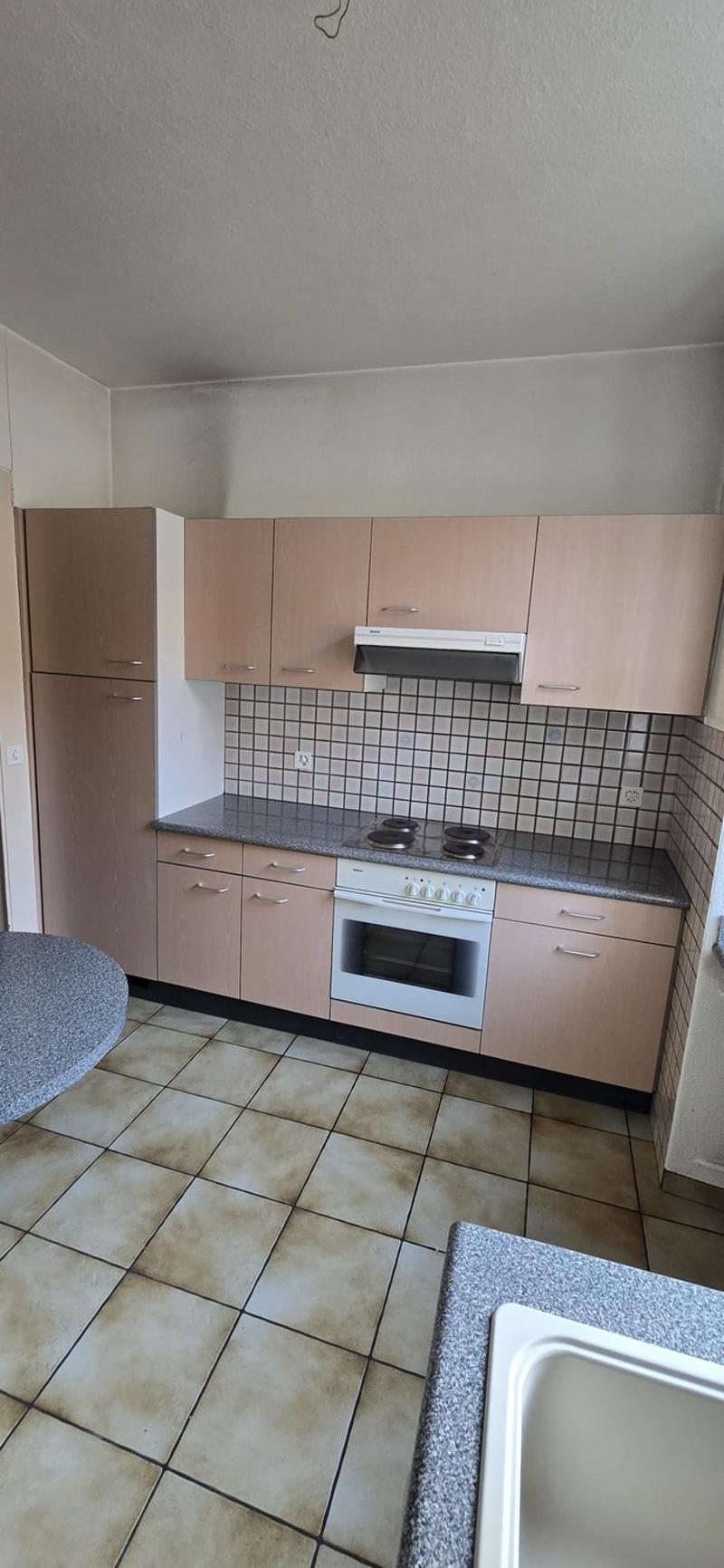 The image shows a kitchen with beige cabinets, a tiled backsplash, a white stove, and a dishwasher. The floor is tiled with a light-colored tile.