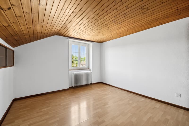 This image shows a room with a wooden ceiling and hardwood flooring. The room has white walls and a large window that provides natural light. There is a radiator visible in the corner of the room.