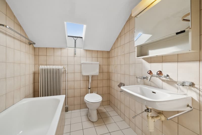 Tiled bathroom with a bathtub, toilet, and sink. The room has a skylight and radiator. The walls are covered in beige tiles.