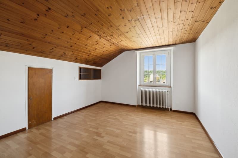 This image shows a room with a wooden ceiling and hardwood floors. The room has a large window that provides natural light and a view of the outdoors. There is a wooden door and a built-in shelving unit on the wall. The room appears to be empty and ready