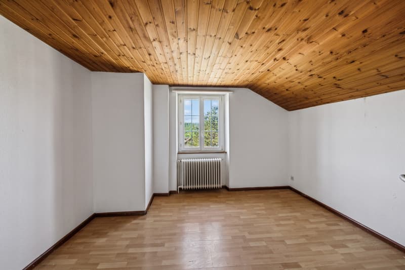 This image shows a room with a slanted wooden ceiling, white walls, and a large window overlooking a scenic outdoor view. The room has a hardwood floor and a radiator heating unit. The space appears to be a bedroom or living area with a simple, rustic des