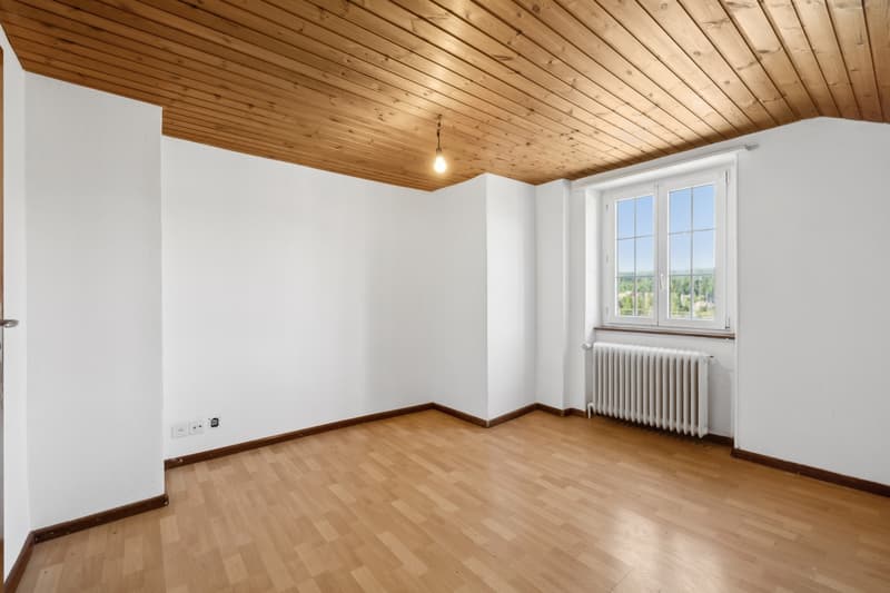 This image shows a spacious room with a wooden ceiling and hardwood floors. The room has large windows that provide natural light and a view of the outdoors. The walls are painted white, creating a bright and airy atmosphere. There is a radiator visible i