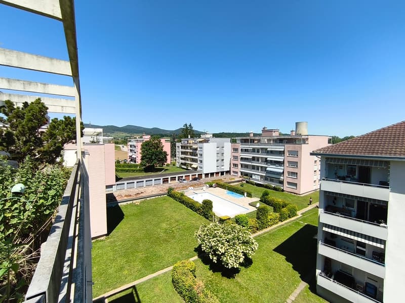 The image shows a multi-story apartment complex with a well-maintained garden and a swimming pool in the center. The buildings have a pink exterior and are surrounded by lush greenery. There are balconies visible on some of the apartments.