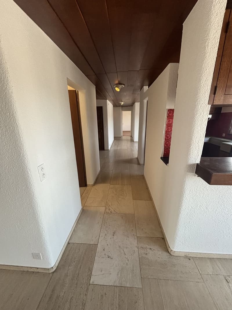 The image shows a long hallway with a tiled floor and white walls. The ceiling has a dark brown paneled design. There are several doors leading off the hallway, and the lighting fixtures are visible along the ceiling.