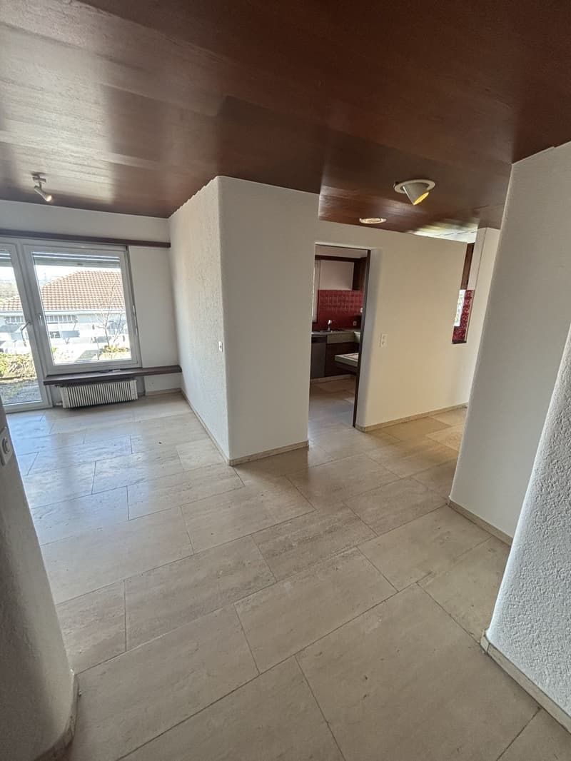 The image shows a spacious living room with a tiled floor, white walls, and recessed lighting fixtures on the ceiling. There is a doorway leading to another room, possibly a kitchen or bedroom. The room appears to be well-lit and has a clean, minimalist a