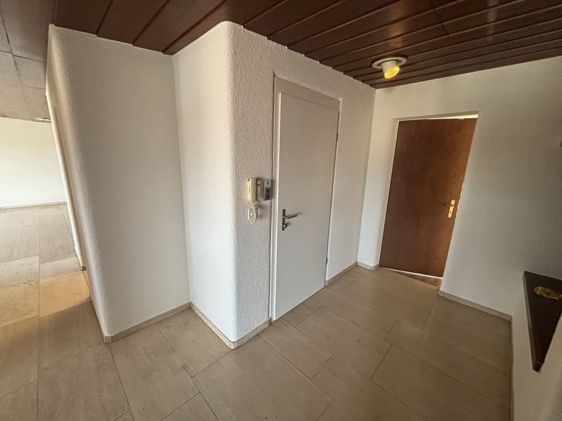 The image shows the interior of an apartment. The entrance features a white door and a wooden ceiling with exposed beams. The floor is tiled, and there is a second door leading to another room. The walls are painted in a light color, creating a bright and