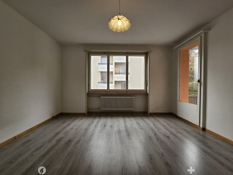 Spacious living room with hardwood flooring, large windows, and a decorative pendant light fixture. The room appears to be in a residential apartment or condominium setting.