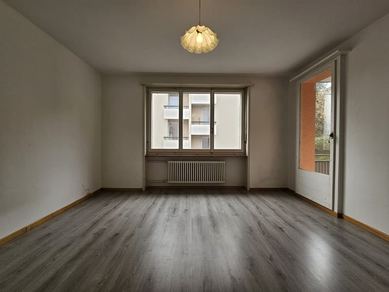 Spacious living room with hardwood flooring, large windows, and a decorative pendant light fixture. The room appears to be in a residential apartment or condominium setting.