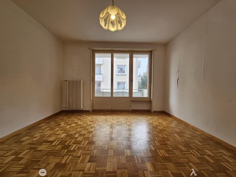 This image shows a spacious room with hardwood parquet flooring, a large window with a radiator underneath, and a decorative light fixture on the ceiling. The walls are painted a neutral color, creating a warm and inviting atmosphere.