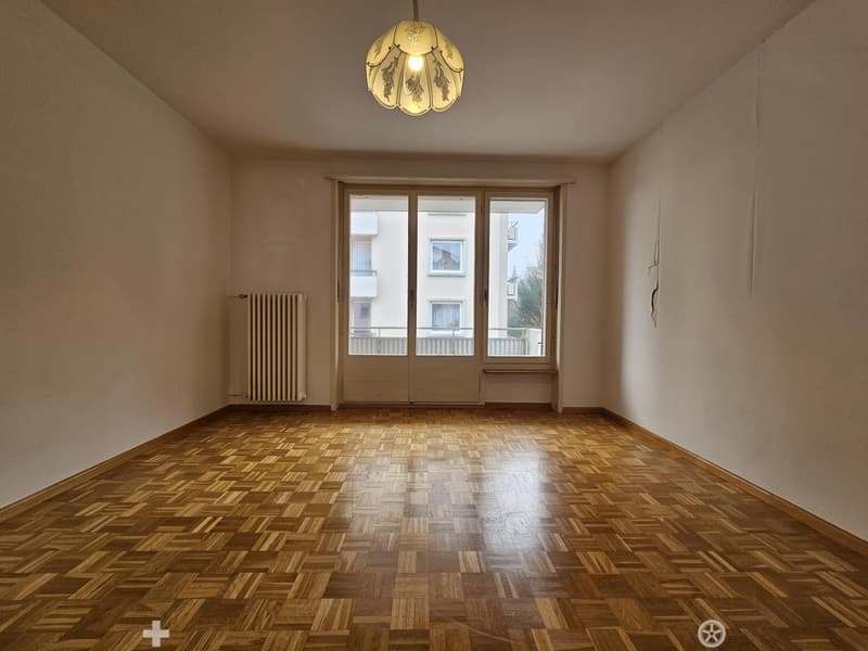 This image shows a spacious room with hardwood parquet flooring, a large window with a radiator underneath, and a decorative light fixture on the ceiling. The walls are painted a neutral color, creating a warm and inviting atmosphere.