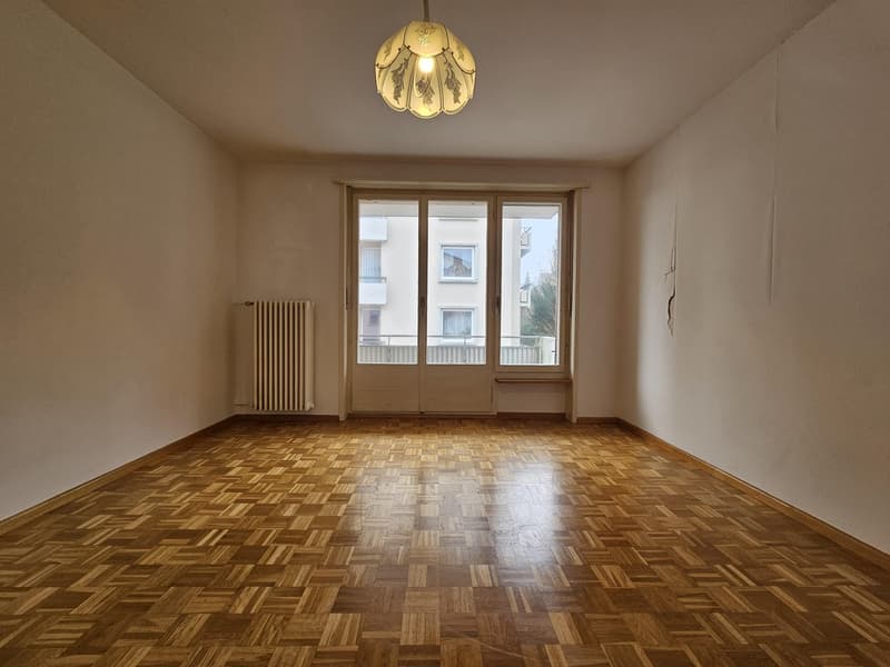 This image shows a spacious room with hardwood parquet flooring, a large window with a radiator underneath, and a decorative light fixture on the ceiling. The walls are painted a neutral color, creating a warm and inviting atmosphere.