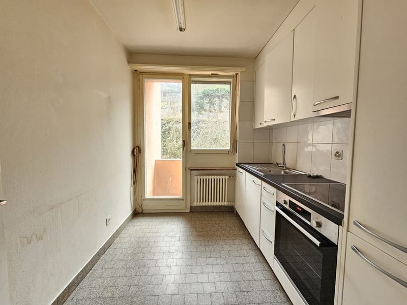 The image shows a small kitchen with a window, tile flooring, white cabinets, and a stove/oven unit. The kitchen appears to be in a residential setting with a view of the outdoors through the window.