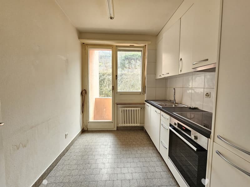 The image shows a small kitchen with a window, tile flooring, white cabinets, and a stove/oven unit. The kitchen appears to be in a residential setting with a view of the outdoors through the window.