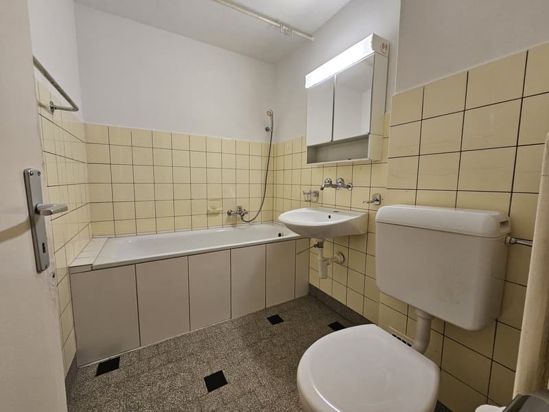 Tiled bathroom with bathtub, sink, and toilet. The walls are covered in beige tiles and there is a mirrored cabinet above the sink.