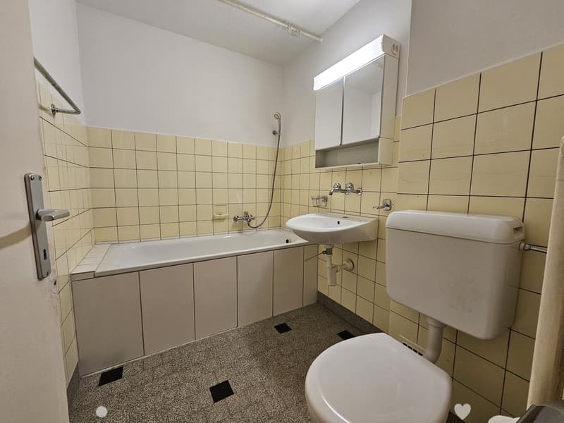 Tiled bathroom with bathtub, sink, and toilet. The walls are covered in beige tiles and there is a mirrored cabinet above the sink.